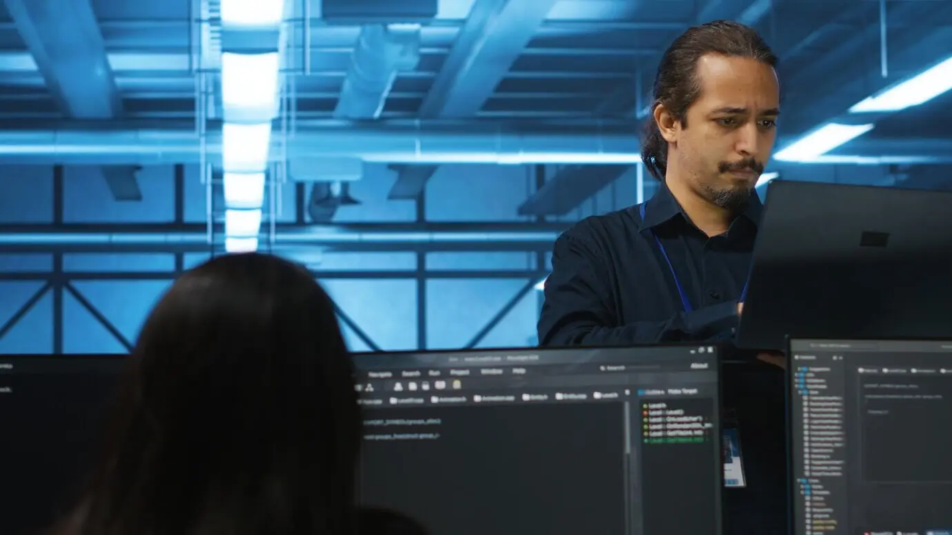 Porträt eines lächelnden Managers, der einen Laptop benutzt und ein Team im Serverraum beaufsichtigt.